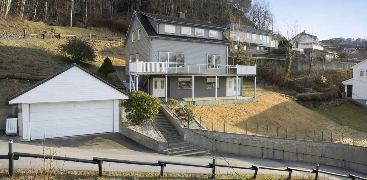 Eiendommen ligger i skrått terreng med frittstående garasje i forkant, steintrapp opp til inngangspartiet og en langstrakt balkong.