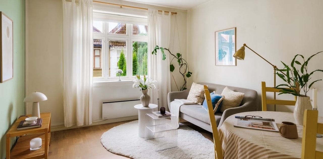 Stuen har tregulv, en grønn fondvegg og et stort vindu som slipper inn rikelig med dagslys mot sofaen og spiseplassen.