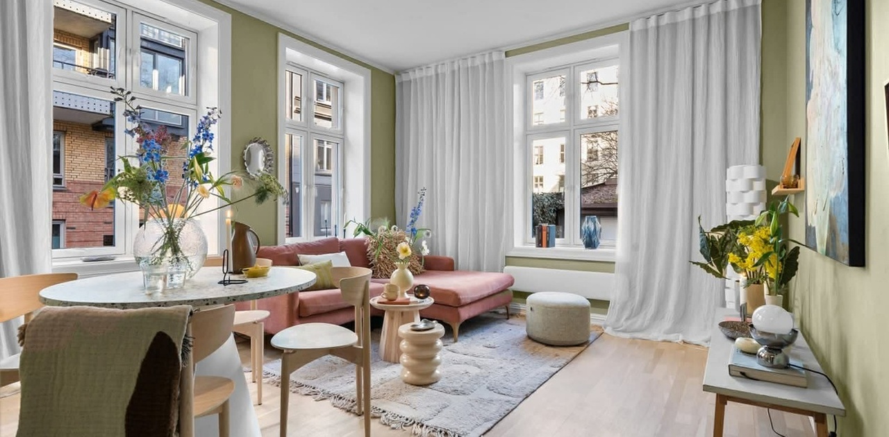 Stuen har olivengrønne vegger, lys parkett og store vindusflater med gulvlange gardiner inn mot en stilren sittegruppe og spiseplass.